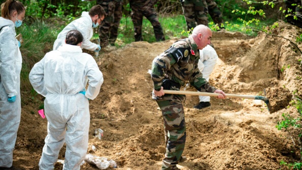 Un gendarme en treillis creuse la terre du charnier avec 4 gendarmes en combinaisons blanches qui exhument des corps et cherchent des preuves.