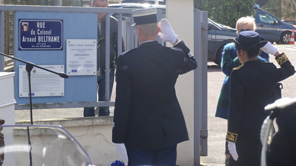 L'autorité militaire et l'autorité civile, de dos, au garde-à-vous, devant la gerbe de fleurs déposée sous la plaque de rue portant le nom du colonel Arnaud Beltrame.