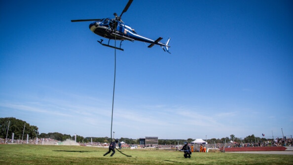 Un hélicoptère de la gendarmerie et deux militaires glissant le long de la corde sur de l'herbe. AU fond, le public