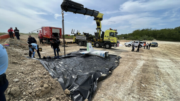La queue du fuselage d'un avion posé sur une grande bâche plastique  noire, devant un camion grue jaune et un camion rouge. Divers enquêteurs travaillent sur la zone dont plueisuers gendarmes.