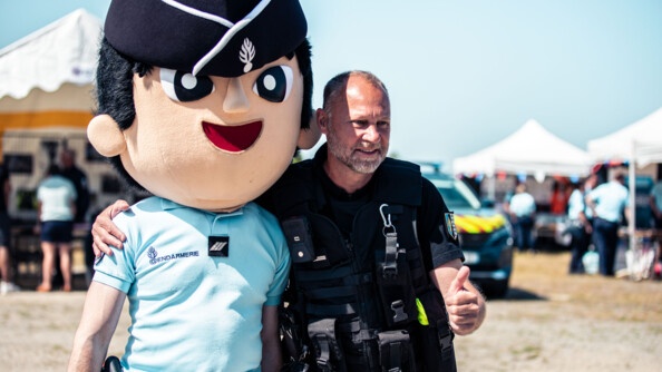 Un gendarme pose avec un autre gendarme qui porte la tête énorme de la mascotte, le gendarme Théo.