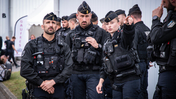 Trois gendarmes mobiles de face dont un lieutenant féminin qui montre quelque chose au loin de son bras gauche. Derrière eux d'autres gendarmes mobiles