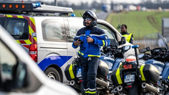 Un motard de la gendarmerie marche. Derrière lui, deux motos ainsi qu'un véhicule de la police. En amorce devant, un autre véhicule blanc et flou.