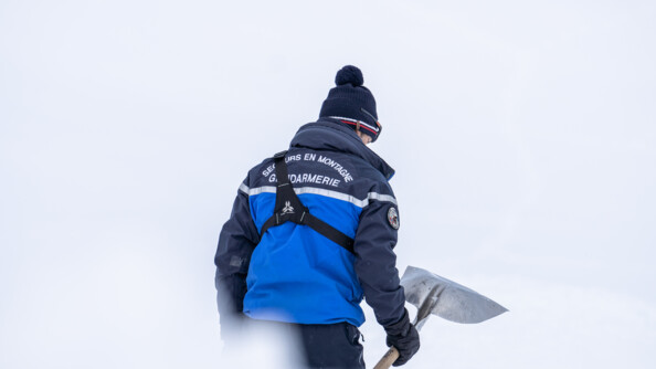 Dans un paysage enneigé flouté, un homme se tient debout, de dos, vêtu d'une tenue de ski bleue. il tient une large pelle métallique dans sa main droite.