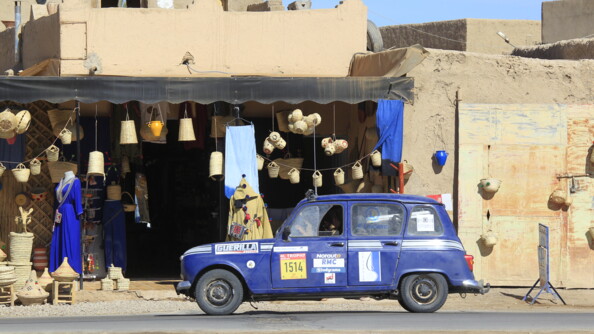 Une Renaud 4L bleu marine, couverte d'enseignes diverses sur les portières, est stationnée devant une boutique marociane d'objets artisanaux (panier en osier et en faïence, robe bleue, pantalon bleu...)
