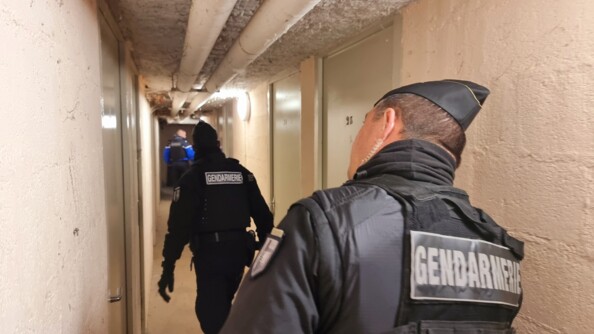 Deux gendarmes mobiles et un gendarme départemental dans un couloir blanc des parties communes au sous-sol d'un immeuble