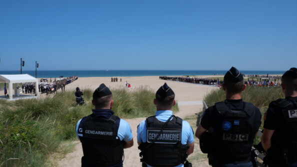 Quatre gendarmes de dos au premier plan surveillent la cérémonie sur la plage, avec la mer au fond.