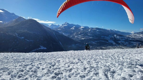 Un parapente décolle sur un sol enneigé, dans un paysage montagneux. Le ciel est bleu et le paysage est ensoleillé