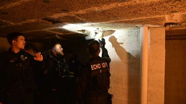 Trois gendarmes dans la pénombre d'une cave ; l'un d'eux projette la lumière de sa lampe vers une cavité dans le plafond, dans laquelle un deuxième militaire regarde.