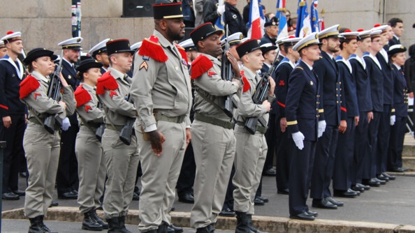 Des militaires, certains en tenue beige, d'autre en tenue bleue marine, se tiennent debout alignés dans la rue. Ils sont postionnés de biais par rapport au photographe. Certains portent un fusil serré contre leur poitrine. On aperçoit plusiers drapeau derrière eux.