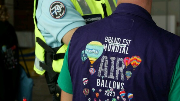 Au premier plan, un personnel de l'événement avec un polo avec pour inscription "Grand Est Mondial Air Ballons 2023 France". Autour de cette inscription des ballons. Au second plan, un gendarme de la GTA