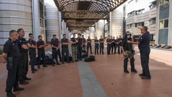 Les stagaires sont en train d'écouter un cadre italien qui montre l'équipement d'un gendarme lors d'une mission de maintien de l'ordre. Ils sont dehors, dans l'école, dans une rue pavée.