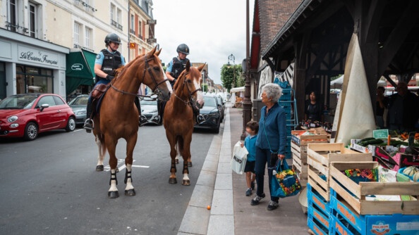 A gauche de l'image, dans une rue, deux chevaux montés par des gendarmes, lesquels sont en train de discuter avec une femme agée et un enfant faisant le marché. A droite, le marché couvert