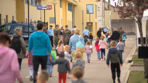 Une foule d'adultes et de jeunes enfants avancent dans une rue. sur leur gauche se trouve un bâtiment jaune. on aperçoit un panneau de gendarmerie, un gendarme et une voiture de gendarme devant le bâtiment