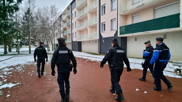 Trois gendarmes mobiles et deux gendarmes départementaux, de dos, marchent en avant le long d'une résidence