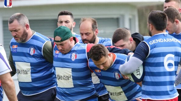 La première et deuxième ligne d'une équipe de rugby, au maillot bleu, se prépare à la mếlée. A droite, un joueur de dos tient le ballon sous son bras gauche