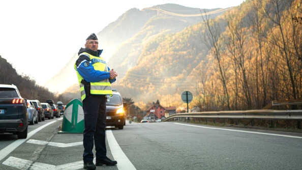Un gendarme avec une chasuble jaune  vérifie l'état du trafic sur la route. A gauche on voit une colonne de voitures. Au fond, les montagnes.
