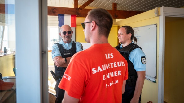 Deux gendarmes, un homme et une femme, échangent avec un sauveteur en mer (de dos avec un tee-shirt rouge).