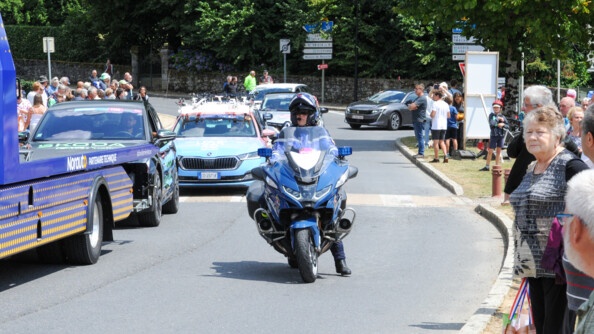 Un motocycliste gendarmerie prêt à mettre pied-à-terre au centre de l'image; de part et d'autre de la route des spectateurs ; à l'arrière de la moto plusieurs véhicules