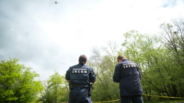 Deux gendarmes de l'Institut de recherche criminelle de la gendarmerie nationale sont dans une clairière. Ils utilisent un drone pour faire une reconnaissance de zone.