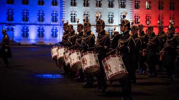 La musique de la garde republicaine, vue de profil gauche, avec 6 tambours alignés et les trompettes alignées au second rang. EN fond, l'etat major de l'EOGN illuminé en bleu blanc rouge