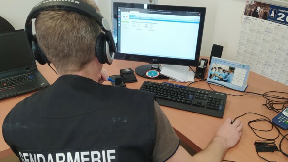 Un gendarme installé face à un ordinateur avec sur le dos, un gilet où est noté "Gendarmerie - Section de Recherches"