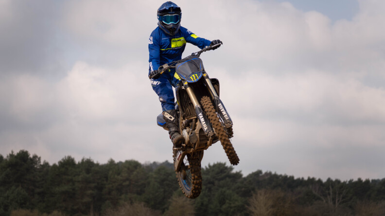 un motard de la gendarmerie vu de trois quart avant gauche et vu en conter-plongé suate dans les airs. En arrière-plan en bas, une forêt de résineux, en haut le ciel gris