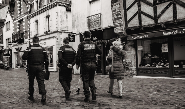 Agrandir l'image 2 gendarmes et un policier de dos ainsi qu'une femme, marchant dans une rue pavée.