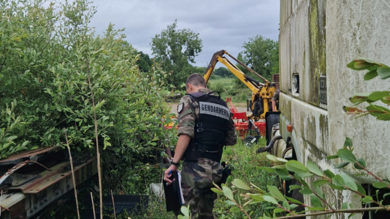 Un gendarme inspecte un terrain, au milieu d'une végération fournie. A sa droite se trouve le mur en béton d'un bâtiment, et plus loin, un engin agricole