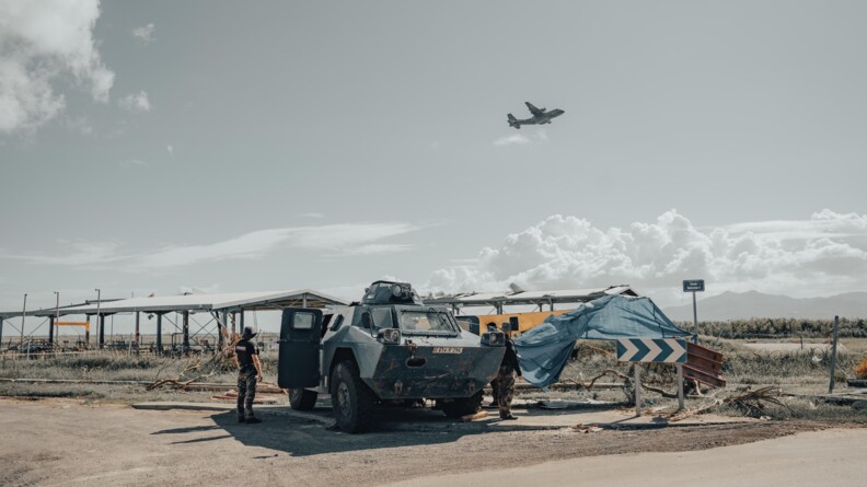 un v&eacute;hicule blind&eacute; bleu avec de aprt et d'autres dux gendarmes devant des hangars en t&ocirc;le fragile. Dans le ciel en haut &agrave; droite, un avion