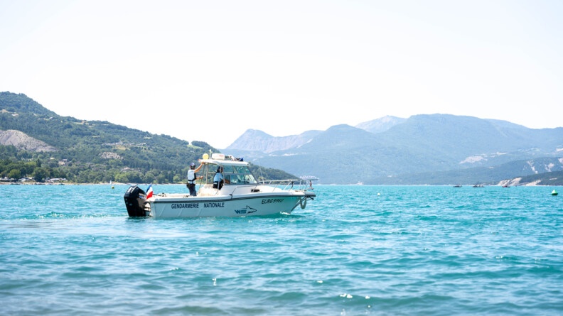 Une navette blanche portant l'insription GENDARMIE NATIONALE en lettres foncées, navigue sur un lac. Deux personnes se tiennent debout à son bord. Le lac est encerclé de reliefs.