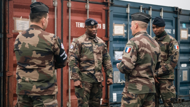 Agrandir l'image De dos, deux prévôts échangent avec deux militaires du train qui sont de face, devant des containers