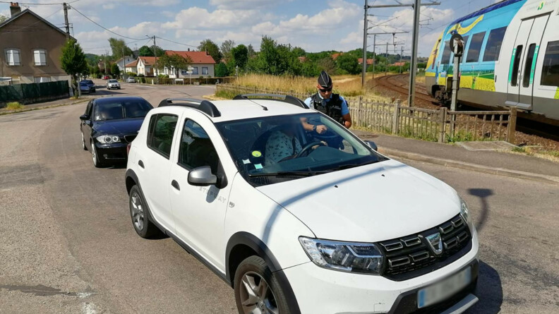 Un gendarme dispense des conseils de pr&eacute;vention au conducteur d'un v&eacute;hicule blanc arr&ecirc;t&eacute; &agrave; un passage &agrave; niveau, tandis qu'un train passe. Au second plan, un second v&eacute;hicule noir.