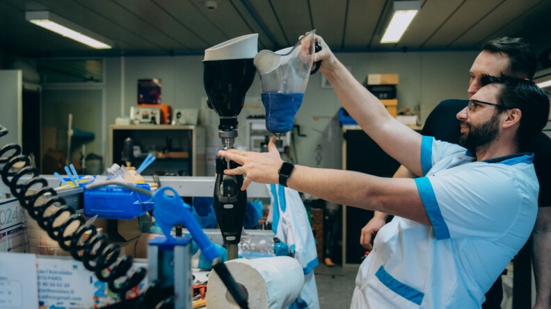 Dans un laboratoire, un homme de profil, portant une blouse blanche, manches courtes, bord&eacute;e de bleu, est debout, observant deux proth&egrave;ses qu'il tient &agrave; bout de bras. Un homme v&eacute;tu d'un pantalon et d'un tee-shirt noirs est &agrave; ses c&ocirc;t&eacute;s.