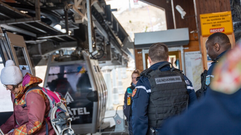 Agrandir l'image Deux gendarmes, se tiennent debout, de dos, à l'endroit où des touristes s'apprêtent à monter dans de petites télécabines.