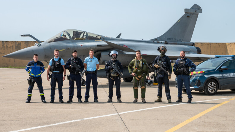 Agrandir l'image Huit militaires en ligne, de gauche à droite, un motard de la gendarmerie, un gendarme départemental, un gendarme de PSIG, un officier de gendarmerie départementale, un membre de l'antenne GIGN d'Orange, un pilote de l'armée de l'Air et de l'Espace (AAE), un autre gendarme du GIGN d'Orange et un pilote d'hélicoptère gendarmerie. EN arrière plan à droite, un 5008 gendarmerie, et en arrière plan au fond, un rafale de l'AAE