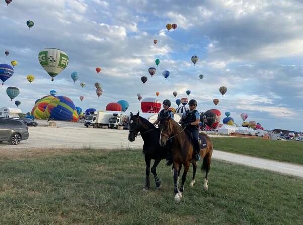 Agrandir l'image Deux cavaliers en patrouille équestre sur une zone herbeuse, à proximité d'un parking, avec un vol de montgolfières en fond.