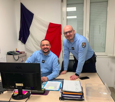 Agrandir l'image Deux gendarmes souriants se tiennent derrière un bureau. L'un 'eux est assis, devant un écan d'ordinateur, tandis que le second se tient de bout à ses côtés, les mains posées sur le bureau, le corps légèrement incliné en direction de son collègue. Derrière eux, sur le mur blanc, est érigé un grand drapeau français.