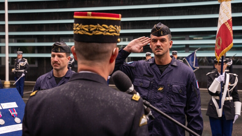 Un gendarme, debout face à l'objectif, effectue un salut militaire en direction d'un haut gradé lui faisant face. Ils sont entourés d'autres militaires. la scène se déroule dans une cour, par temps de pluie.