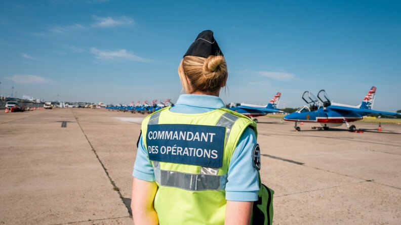 Au milieu de l'image, de dos, une gendarme avec la chasuble jaune siglée "commandant des opérations". Face à elle, le tarmac où sont alignés les avions de la Patrouille de France