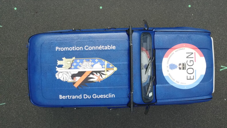 On aper&ccedil;oit vue d'en haut une Renaud 4L bleu marine, couverte d'une grande enseigne sur le toit avec la mention "Promotion Conn&eacute;table Bertrand Du Guesclin", et une seconde sur le capot portant la mention "Ecole des officiers de la gendarmerie nationale EOGN"