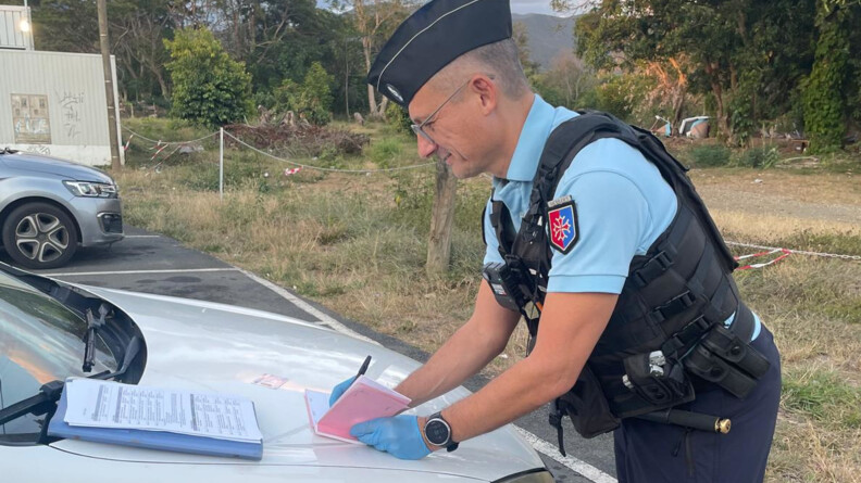 Un gendarme remplit un carnet de verbalisation rose sur le capot d'une voiture blanche. En arrière-plan la nature et un container blanc à gauche
