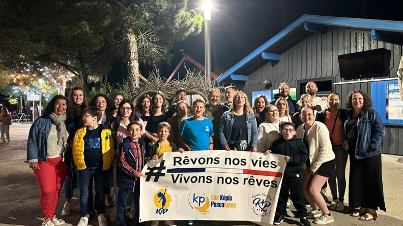 Agrandir l'image Devant un bâtiment en bois au toit bleu à droite, et des arbres à gauche, de nuit, un groupe de 25 personnes dont de nombreux enfants, portant la banière des képis pescalunes