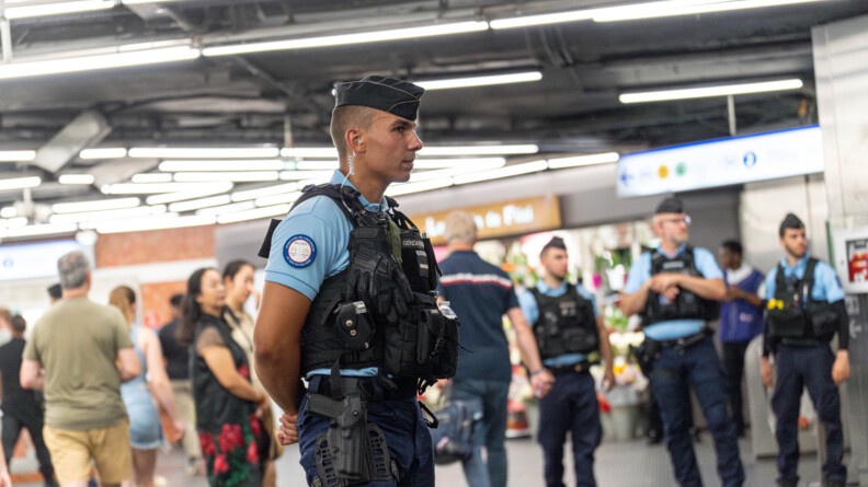Au premier plan, un gendarme départemental de profil, regarde droit devant lui. En arrière plan, trois autres gendarmes de face surveillent le flot de personnes. Au second plan, à gauche, un flot de voyageurs. Dans le fond, une boutique de fleurs.