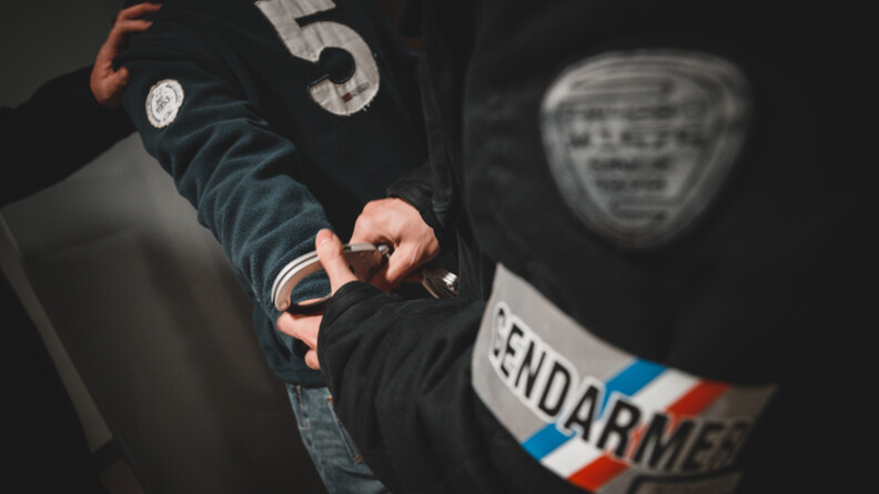 Zoom sur un placement de menottes à un individu portant un haut de survêtement ebleu foncé et un jean, menottes mises par une personne au blouson noir porteur d'un brassard gendarmerie. A gauche de l'image la main d'un tiers tient l'épaule de l'individu menotté