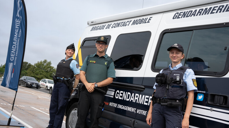A droite, une policière allemande coiffée d'une casquette noire portant la mention en blanc "POLIZEI"  et vêtue d'un pantalon bleu foncé et d'une chemisette bleu clair, recouverte d'un gilet pare-balles bleu foncé. Au milieu, un garde civil espagnol en pantalon noir, t-shirt et casquette vertes. A gauche, une gendarme coiffée d'un calot, portant un pantalon bleu foncé et un polo manches courtes bleu clair, recouvert d'un gilet pare-balles noir. Ils sont debout, côte à côte,  devant un fourgon bleu foncé et blanc, avec des bande jaunes à l'arrière, portant la mention en blanc au centre "Gendarmerie notre engagement votre sécurité (le mot est coupé)" et en haut en bleu foncé sur fond blanc, "Brigade de contact". A gauche, un kakémono bleu portant la mention gendarmerie flotte. On devine que l'on est sur un parking. A gauche en arrière-plan, on voit trois voitures garées.