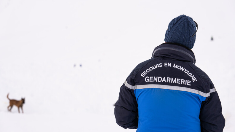 Agrandir l'image Un homme se tient debout, de dos, dans un paysage enneigné, légèrement flouté. il porte un anorak bleu portant dans le dos l'inscription Secours en montagne Gendarmerie. A gauche de l'image, on aperçoit un berger belge malinois, également flouté.