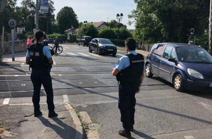 Deux gendarmes vus de dos, au bord de la route, &agrave; proximit&eacute; d'une voie ferr&eacute;e sans barri&egrave;re. En arri&egrave;re-plan, deux v&eacute;hicules passent.