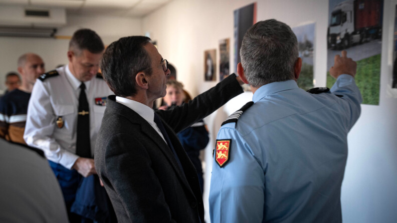 Un colonel de gendarmerie, de trois-quart dos, v&ecirc;tu d'une chemise bleue manches longues, pointe du doigt un mur o&ugrave; sont accroch&eacute;es des photos. A gauche, de profil, en costume sombre, chemise blanche, le ministre de l'Int&eacute;rieur, pointe &eacute;galement son doigt vers le mur. En arri&egrave;re plan, un officier g&eacute;n&eacute;ral de gendarmerie en chemise blanche - cravate, et dans le fond des personnes en civil.