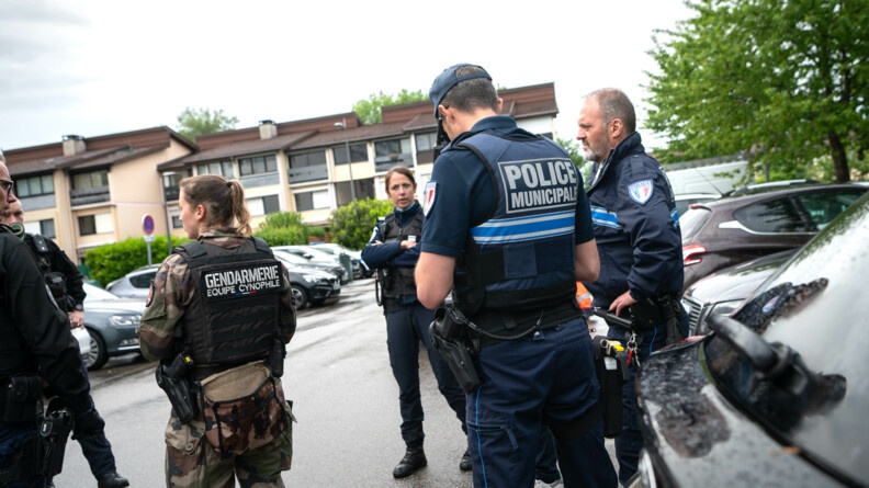 Agrandir l'image Des gendarmes et un policier municipal discutent sur un parking extérieur, entouré d'habitations. le temps est pluvieux.
